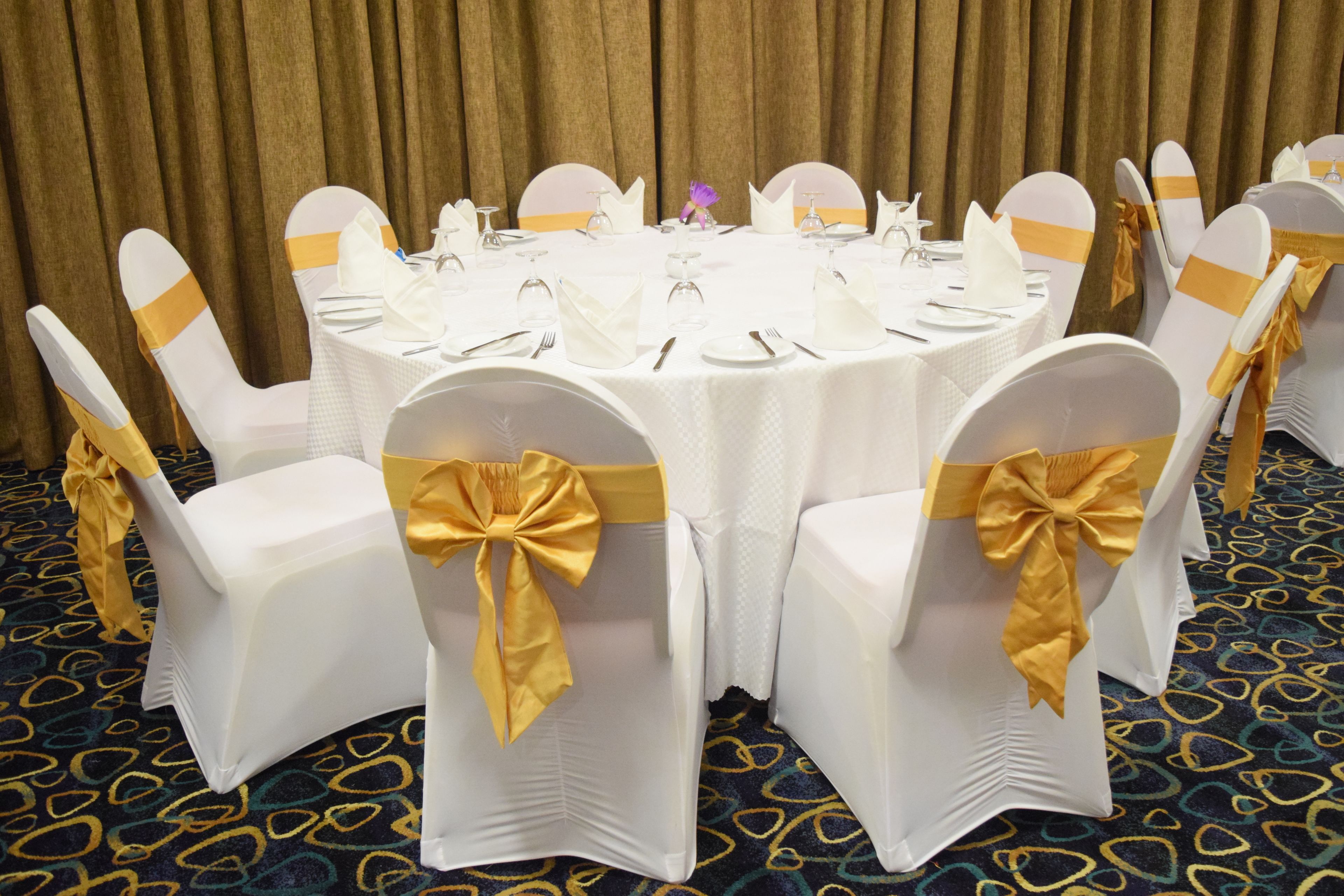A round table with white tablecloths and gold sashes, surrounded by chairs with gold sashes.