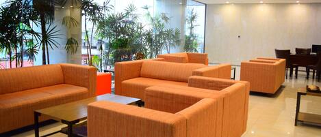 A modern lobby with orange sofas, a wooden coffee table, and large windows with plants.