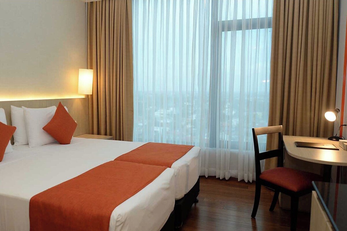 A hotel room with a large bed, two pillows, a desk with a chair, and a window with curtains.
