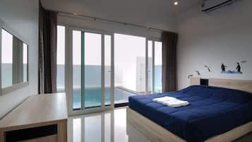 A modern bedroom with a large bed, a wooden desk, and a view of the sea through floor-to-ceiling windows.
