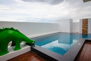 A modern indoor swimming pool with a clear blue tile floor, a wooden deck area, and a green dinosaur-shaped object on the left. - Le Leaf Pool Villa 6 (Hua Hin)