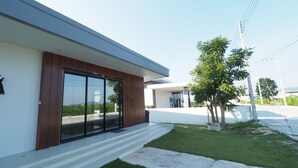 A modern building with a large glass door, a small front yard with grass and a tree, and a clear sky. - Le Leaf Pool Villa 7 (Hua Hin)
