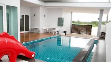 A modern indoor swimming pool with a clear blue tiled floor, surrounded by white walls and a wooden deck area.