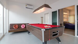 A modern pool table with a red felt surface, a brown cushioned bench with two donut-shaped cushions, and a hanging pendant light. - Le Leaf Pool Villa 7 (Hua Hin)