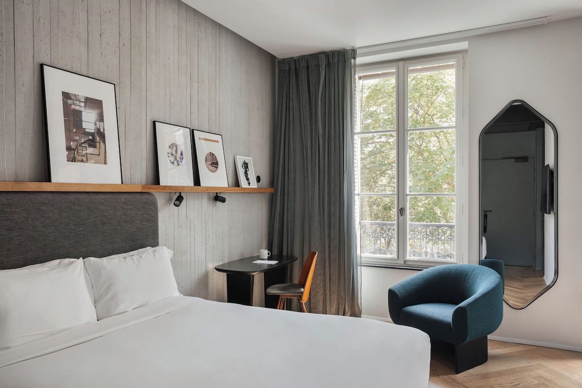 Una habitación de hotel moderna con una cama grande, una mesita con una silla, un ventanal con cortinas y cuadros enmarcados en la pared.