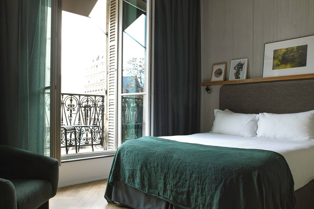 Habitación de hotel con una cama, una silla, una ventana que da a un balcón y un estante en la pared con fotografías.