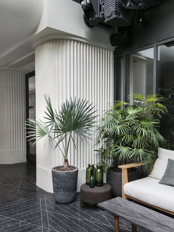 Un intérieur moderne avec une colonne blanche incurvée, un bac à fleurs en pierre contenant un palmier, un banc en bois et un canapé blanc agrémenté d’un coussin.
