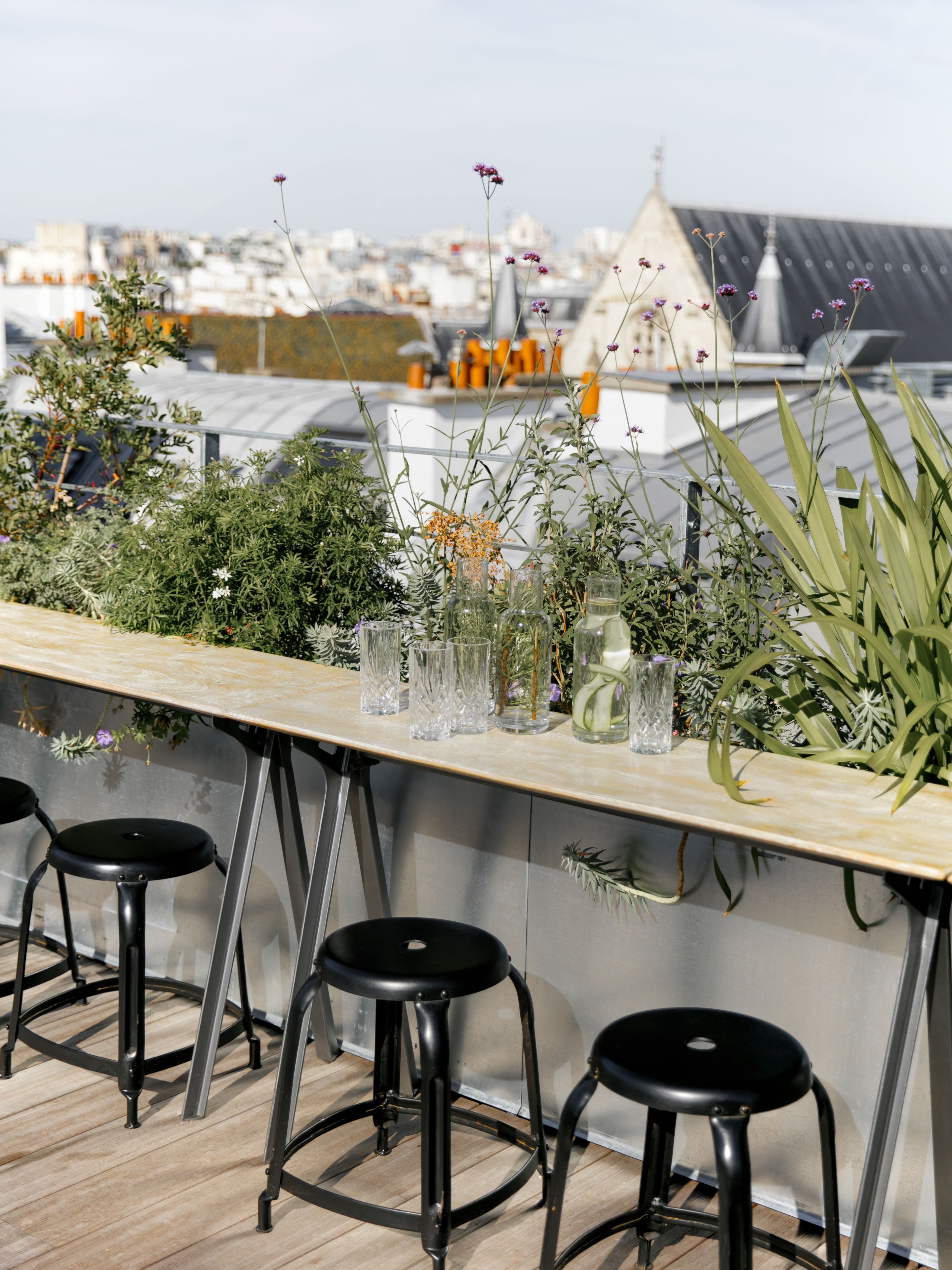 Un bar situé sur le toit, avec une longue table en bois, trois tabourets noirs, ainsi que diverses plantes et différents types de verres.