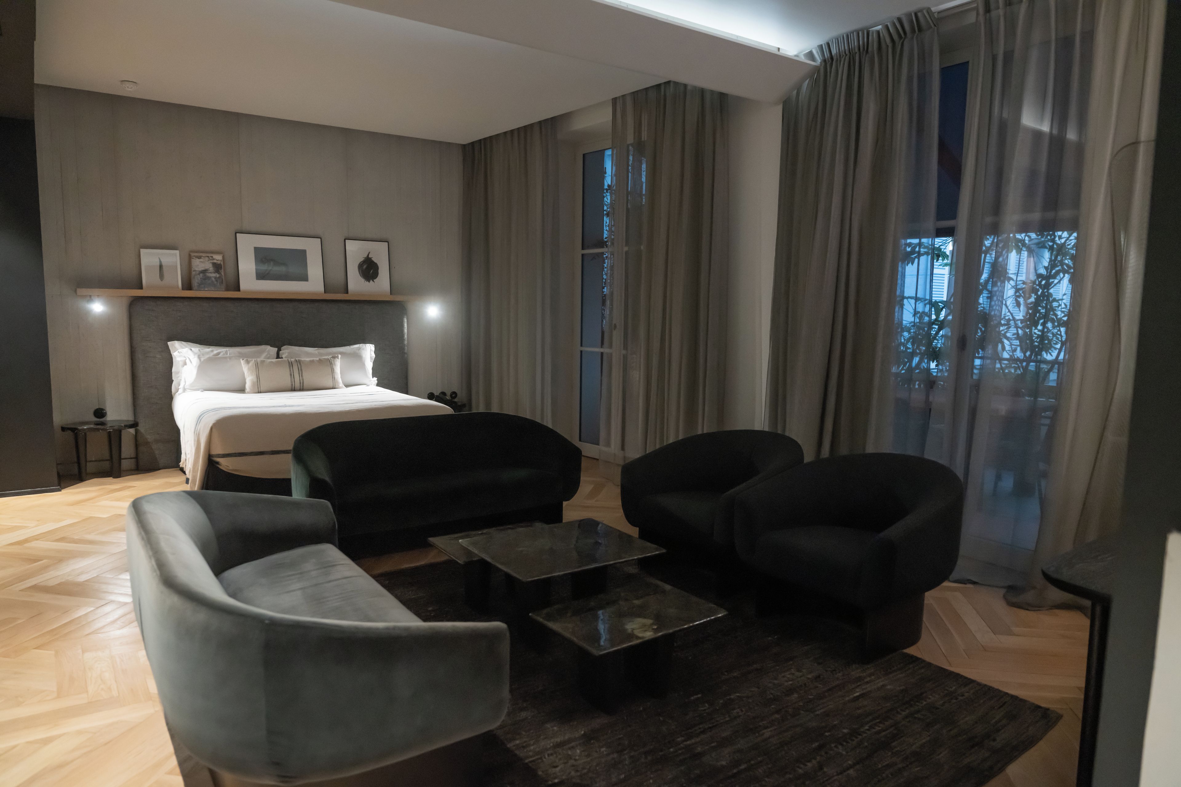 A modern hotel room with a bed, two armchairs, a sofa, a coffee table, and a large window with curtains.