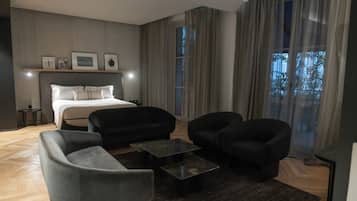 A modern hotel room with a bed, two armchairs, a sofa, a coffee table, and a large window with curtains.