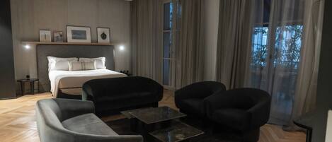 A modern hotel room with a bed, two armchairs, a sofa, a coffee table, and a large window with curtains.