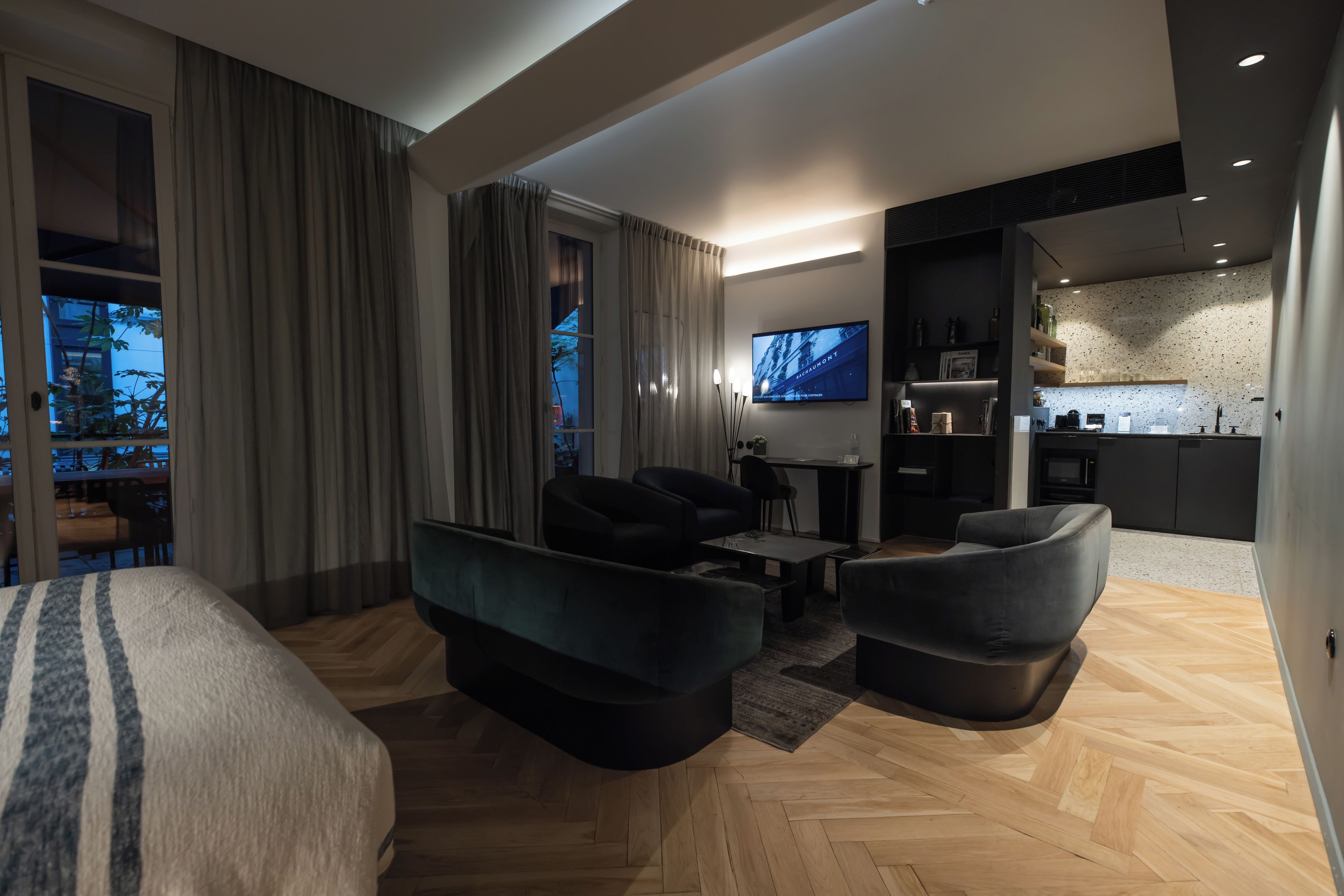 A modern hotel room with a large bed, two sofas, a TV, and a kitchenette.
