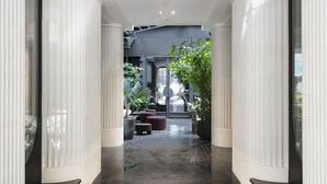 Een moderne hal met donkere betegelde vloer, witte zuilen en een zithoek met planten en een bank.