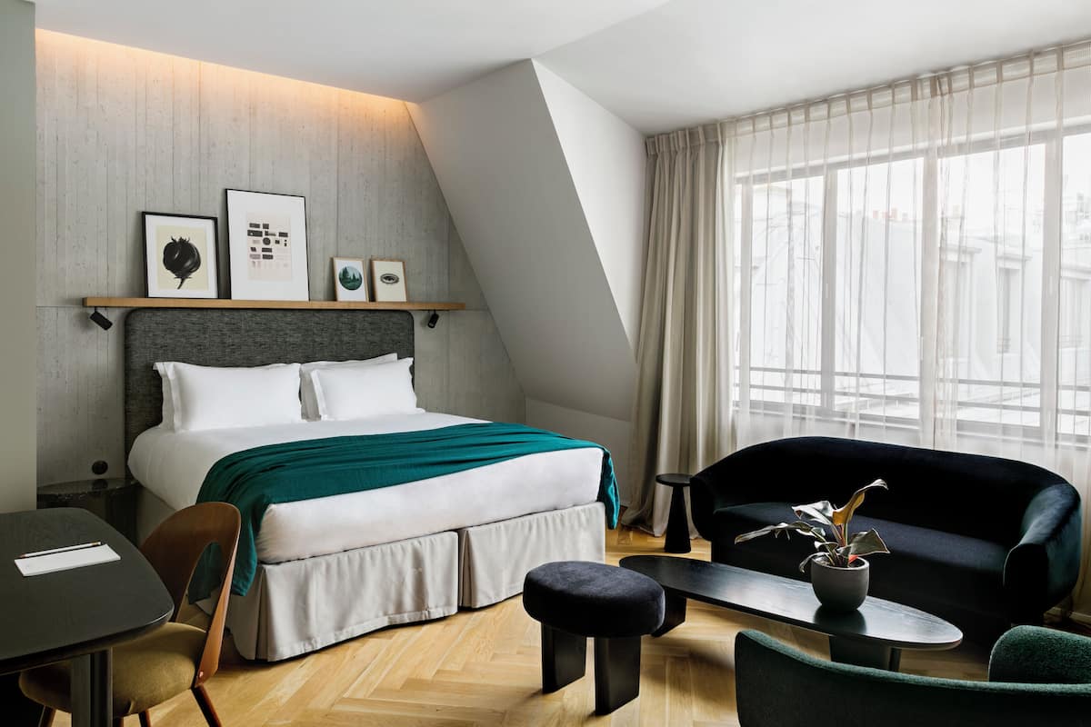 Una habitación de hotel moderna con cama, sofá, una mesita y una silla. Hay cuadros enmarcados en la pared sobre la cama.