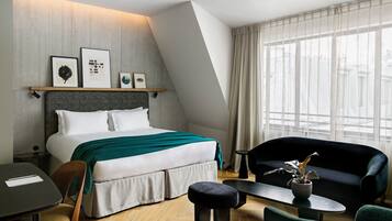 A modern hotel room with a bed, a sofa, a small table, and a chair. There are framed pictures on the wall above the bed.