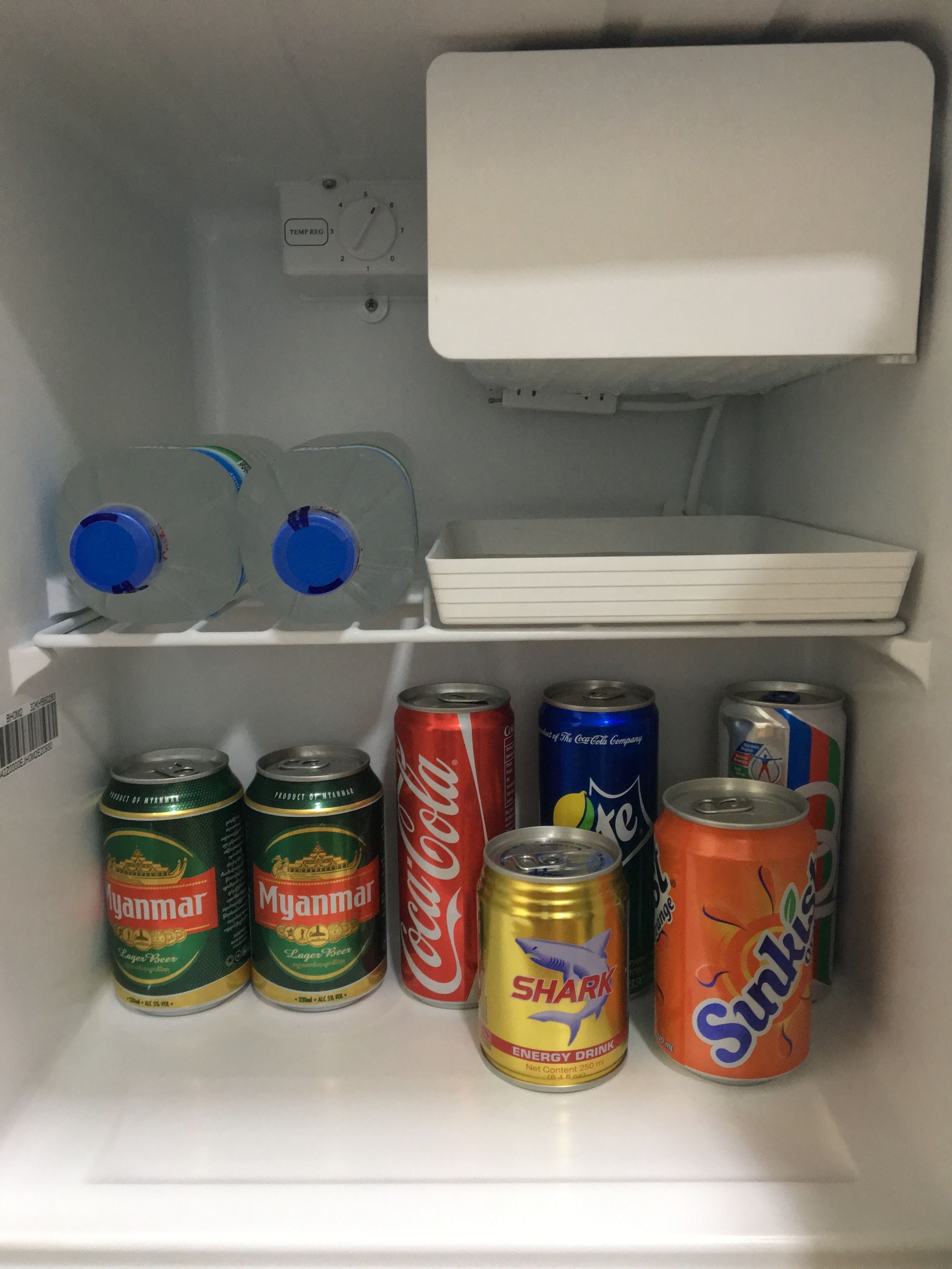 A refrigerator with shelves containing various beverages including cans of Myanmar lager beer, Coca-Cola, and energy drinks.