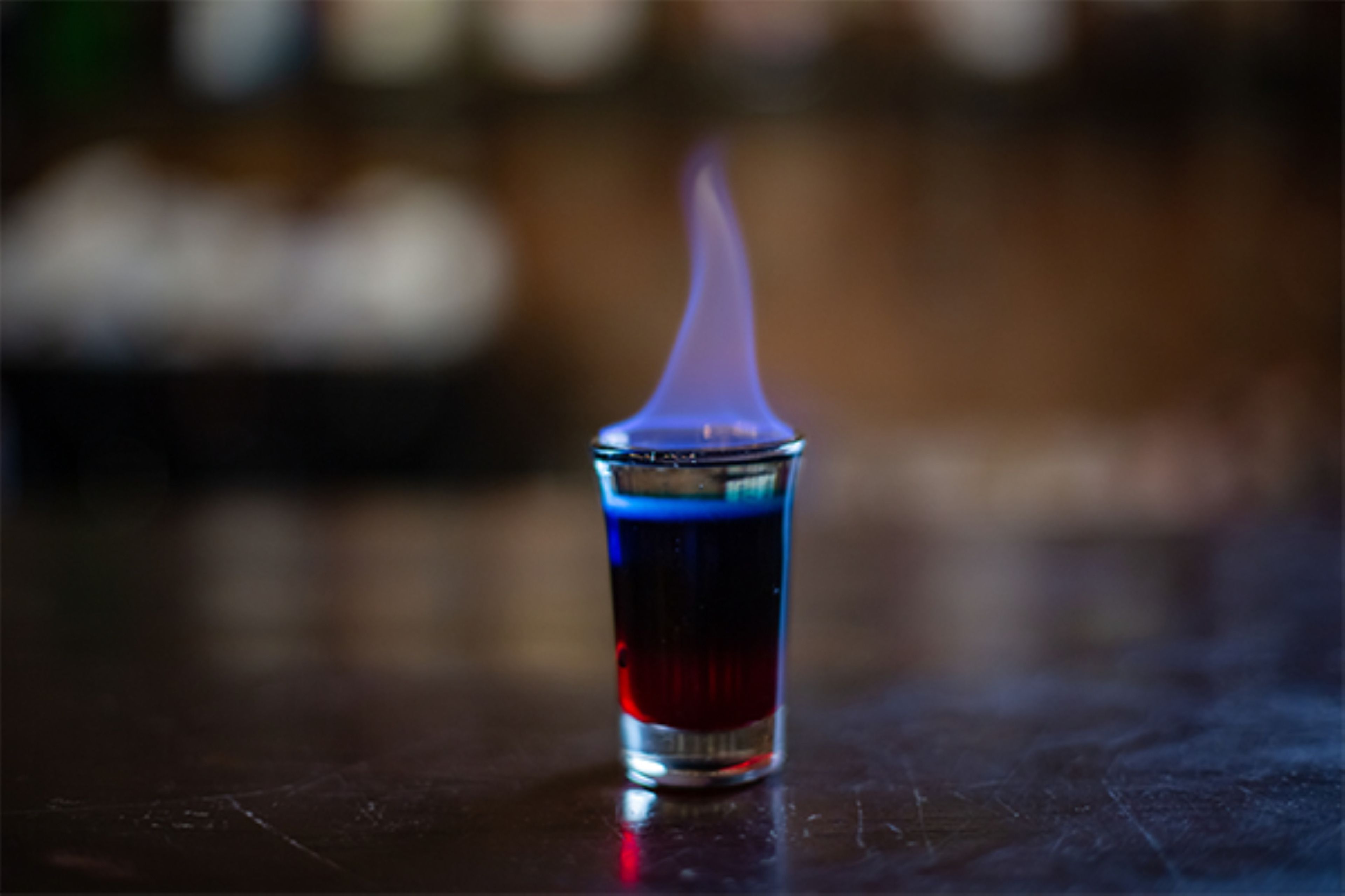 Et nærbillede af et glas med blå væske og en flamme ovenpå, placeret på en reflekterende overflade.