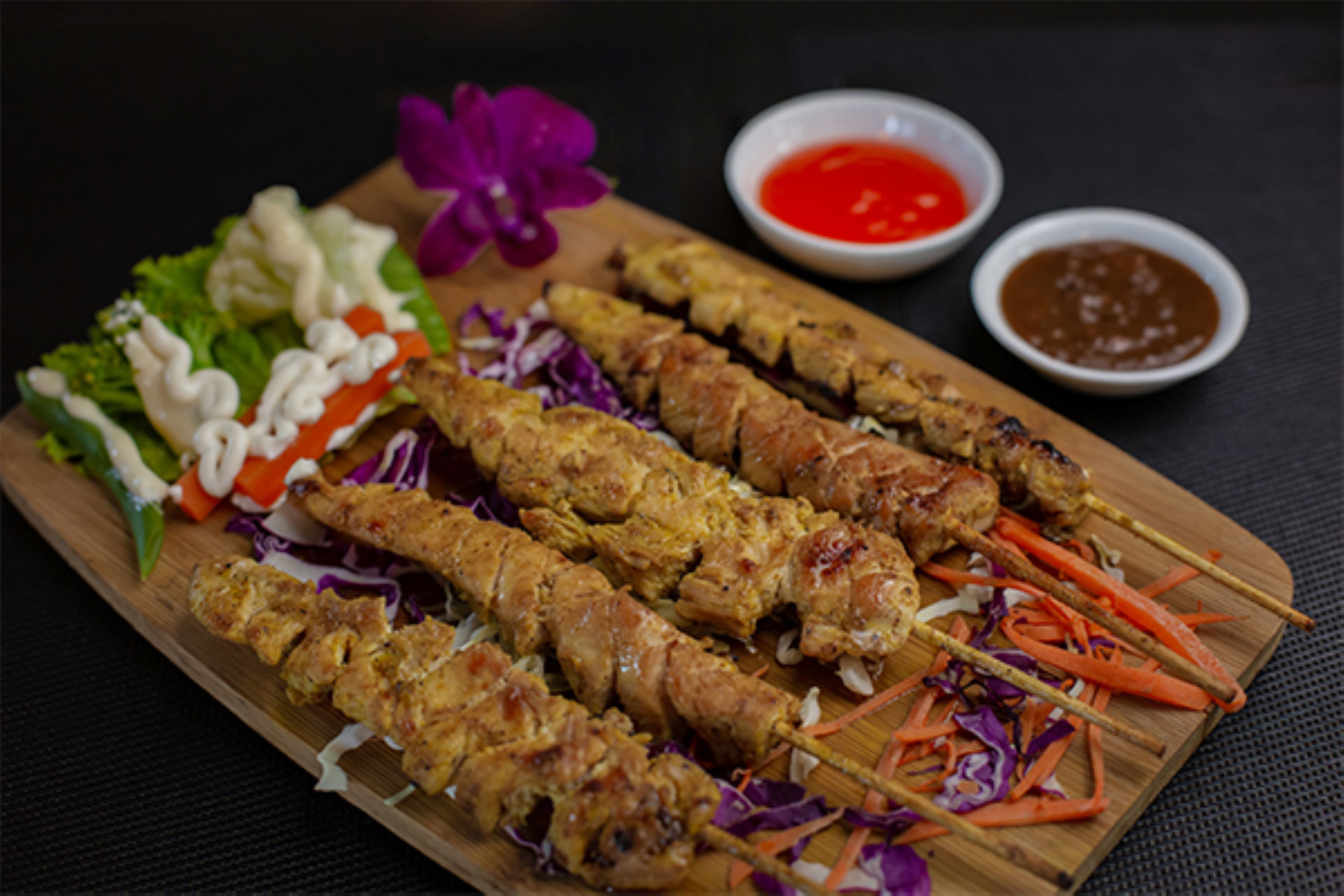 A wooden board with skewered meat, fresh vegetables, and two small bowls of sauce.