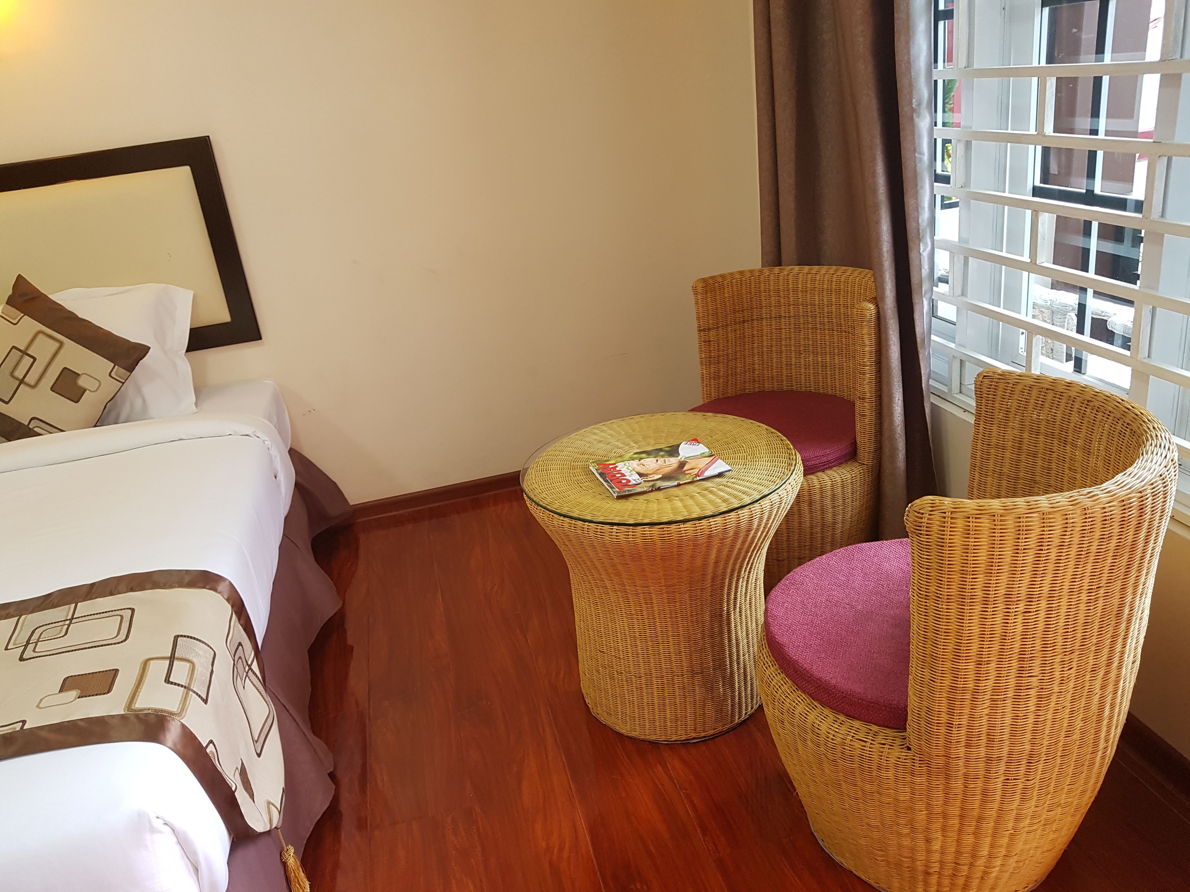 A hotel room with a bed, a wicker chair, a small round table with magazines, and a window with a view of buildings.