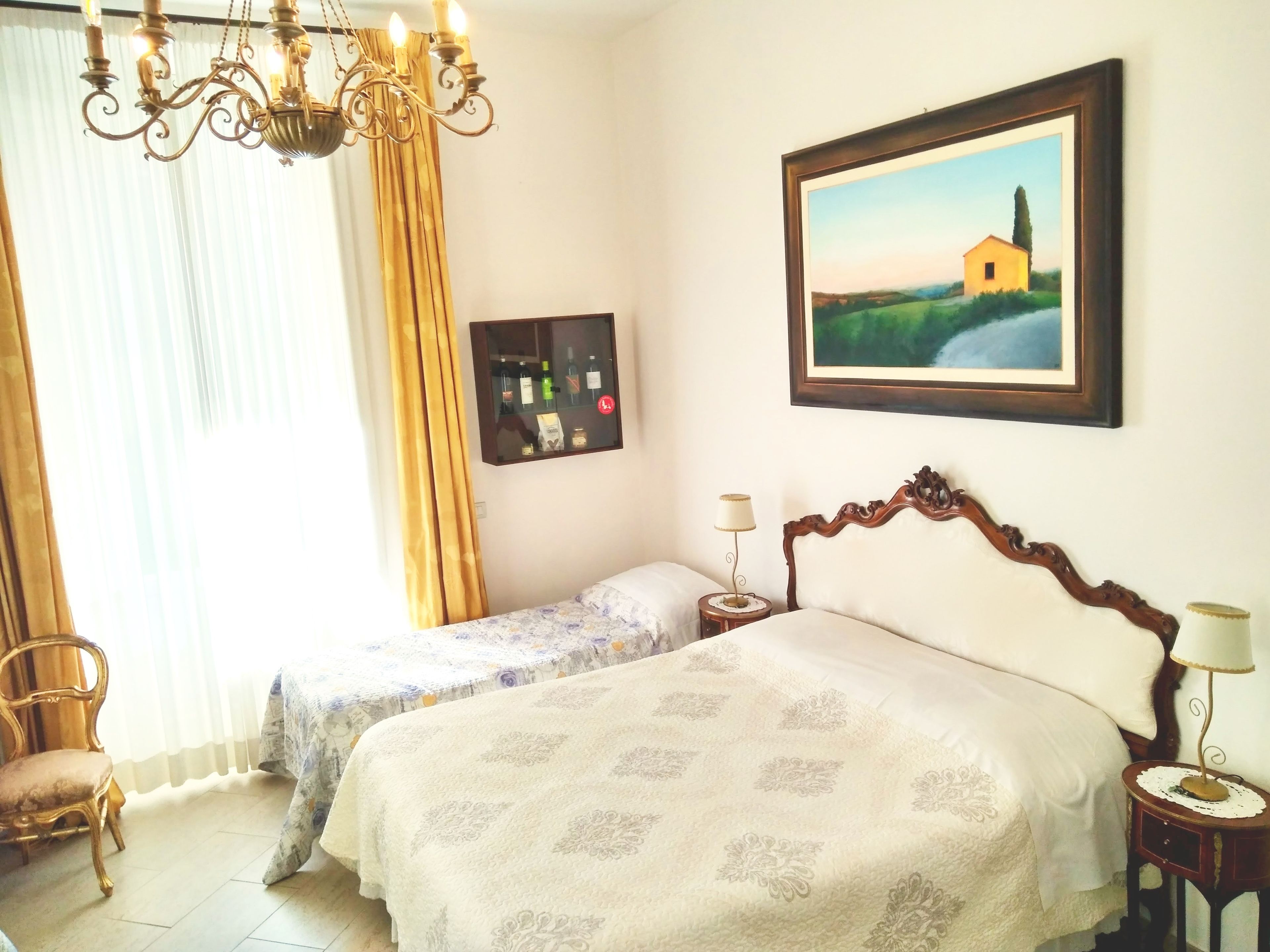 Une chambre d’hôtel avec un grand lit, un lit plus petit, un lustre, un tableau encadré au mur et une table de chevet avec une lampe.