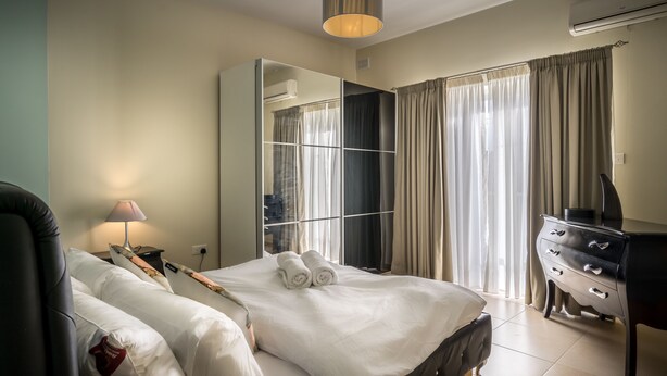 A hotel room with a bed, a desk, a chair, a lamp, a wardrobe, and a large mirror. - Consiglia Apartments - Sliema (Sliema)