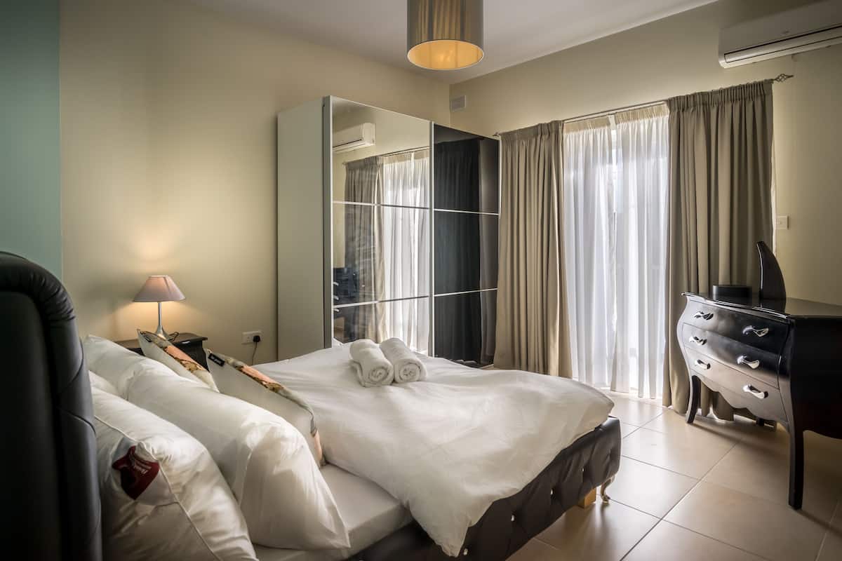 A hotel room with a bed, a desk, a chair, a lamp, a wardrobe, and a large mirror.