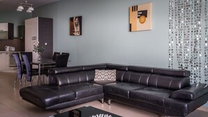 A modern living room with a black leather sectional sofa, a dining area with a table and chairs, and a kitchen in the background. - Consiglia Apartments - Sliema (Sliema)