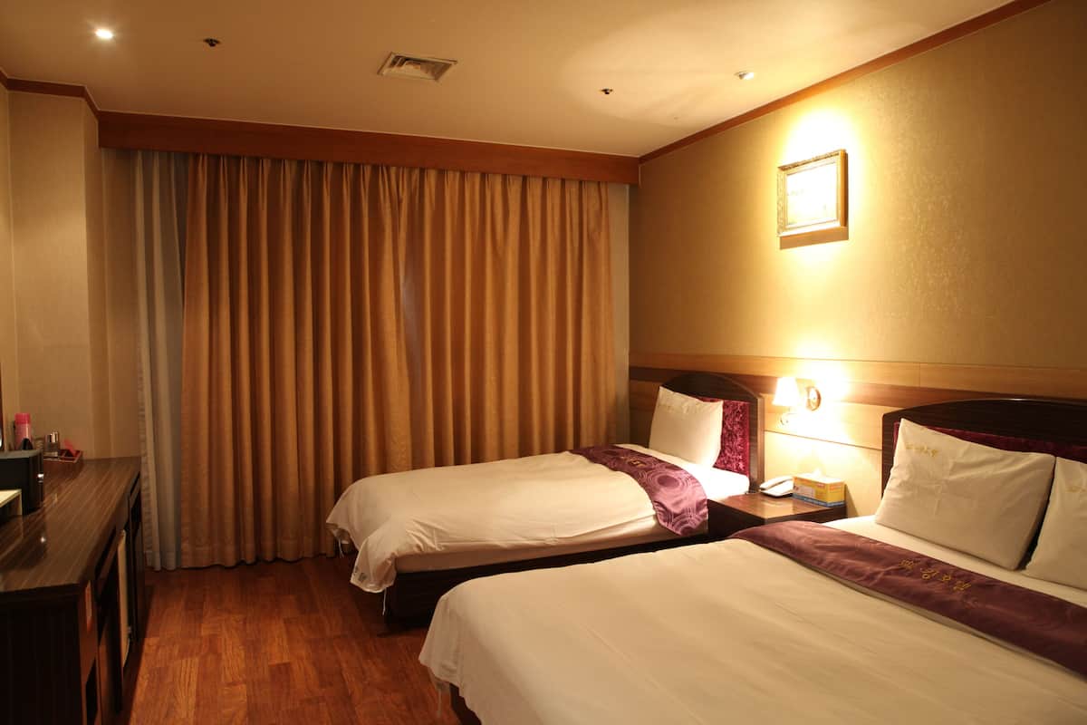 A hotel room with two beds, a wooden desk, a nightstand with a lamp, and a large window with curtains.