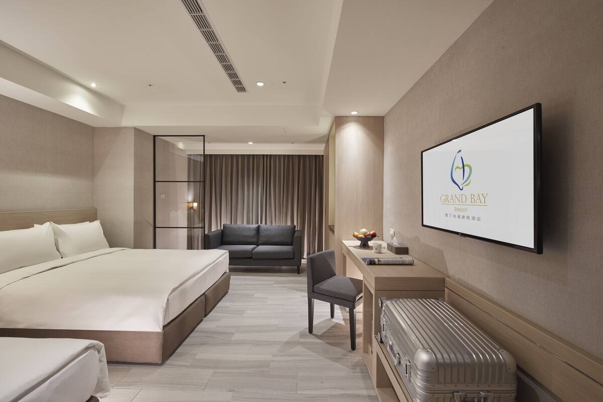 A modern hotel room with a large bed, a desk with a suitcase, and a television displaying the hotel name.