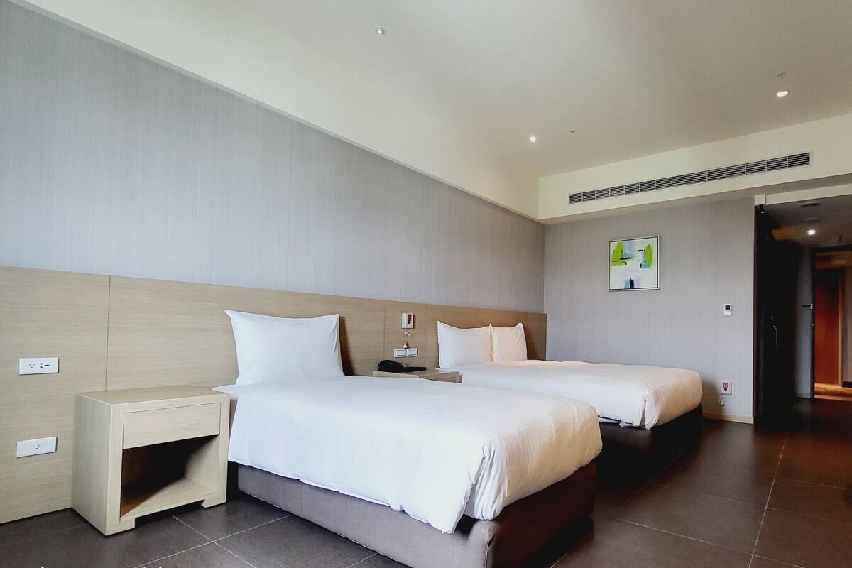 A modern hotel room with two beds, a small bedside table, a wall-mounted TV, and a door leading to another room.