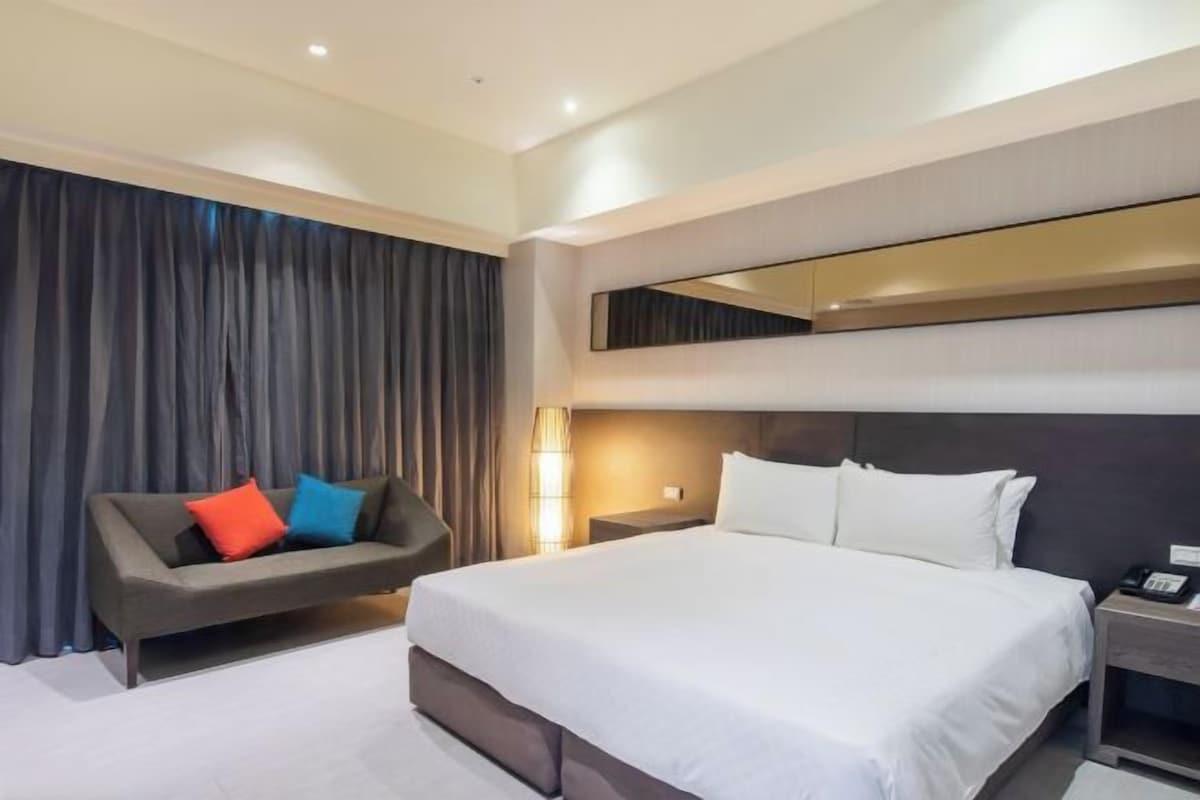 Una habitación de hotel moderna con una cama grande, un sofá con almohadas coloridas, una mesita de noche y un ventanal con cortinas.