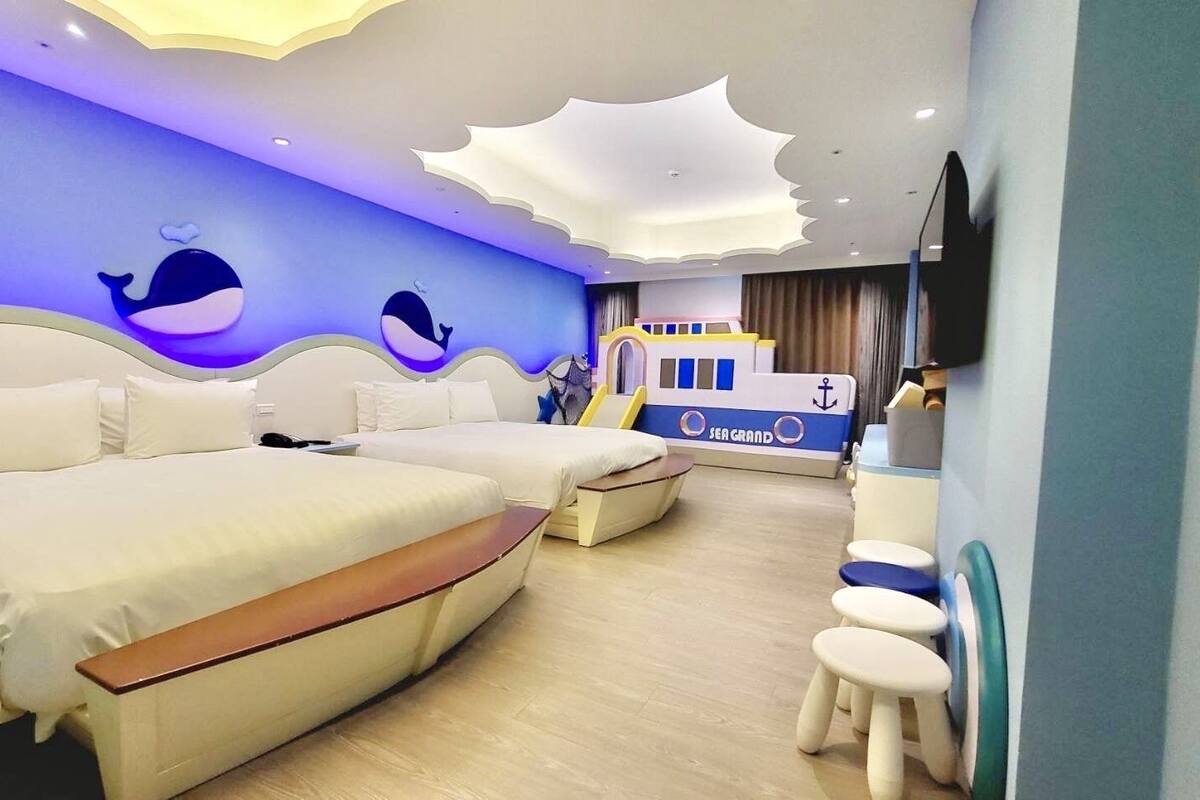 A hotel room with two beds, a small desk, a TV mounted on the wall, and a ship-themed wall decoration.