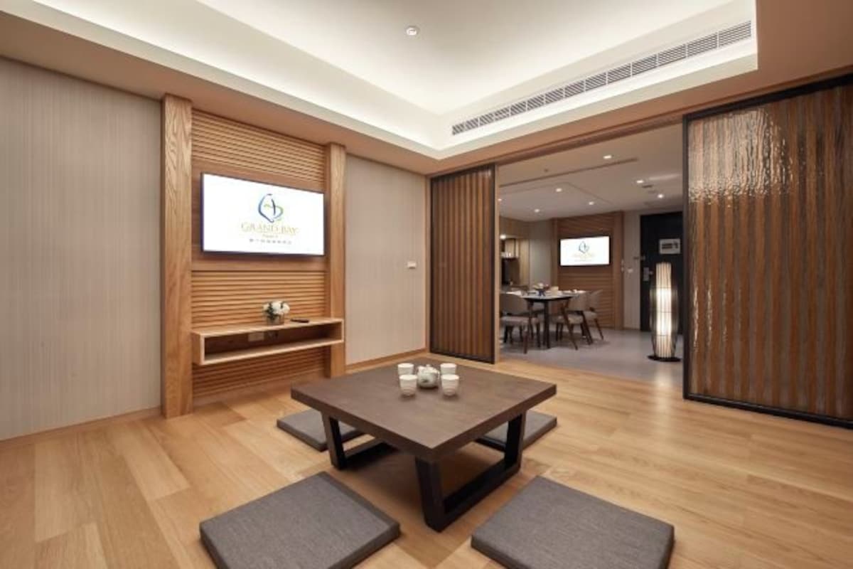Una habitación de hotel moderna con suelo de madera, una mesa central con cuatro sillas, un televisor montado en la pared y una puerta corredera que da a otra habitación.