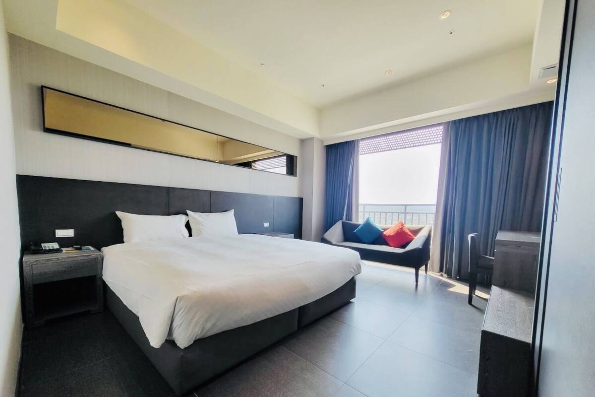 A modern hotel room with a large bed, a desk, and a balcony with a view.