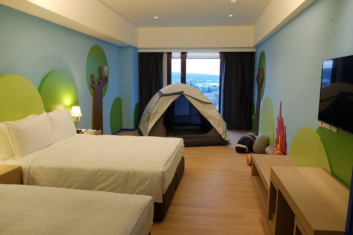 A hotel room with two beds, a tent-like structure, a TV, and a view of the outdoors.