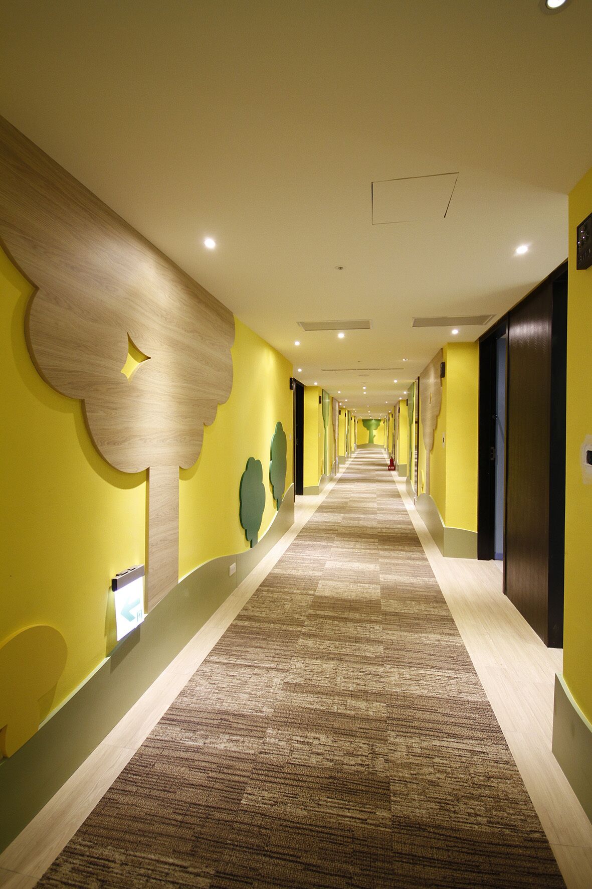 A hotel corridor with a carpeted floor, yellow walls, and a wooden accent wall with a cloud-like design.
