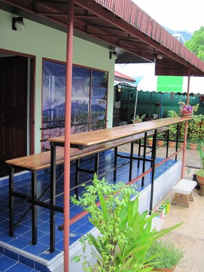 A covered outdoor seating area with a long wooden bench, a tiled floor, and potted plants. - Sakura House Chiangmai (Chiang Mai)