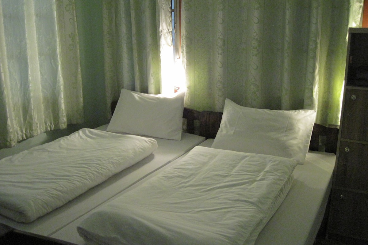 Un lit simple avec du linge de lit blanc et une tête de lit en bois, une table de chevet et un rideau à motifs verts.