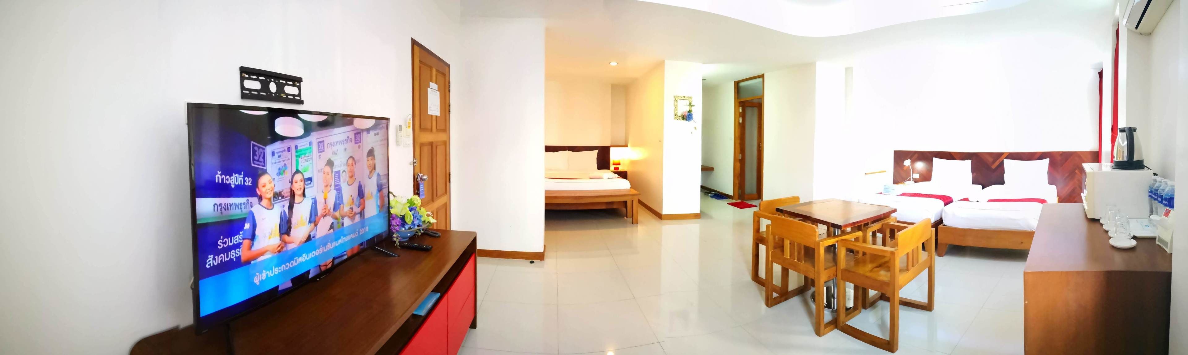 Habitación de hotel con televisión sobre un soporte de madera, cama con ropa blanca, mesa de comedor de madera con sillas y una pequeña cocina con hervidor.