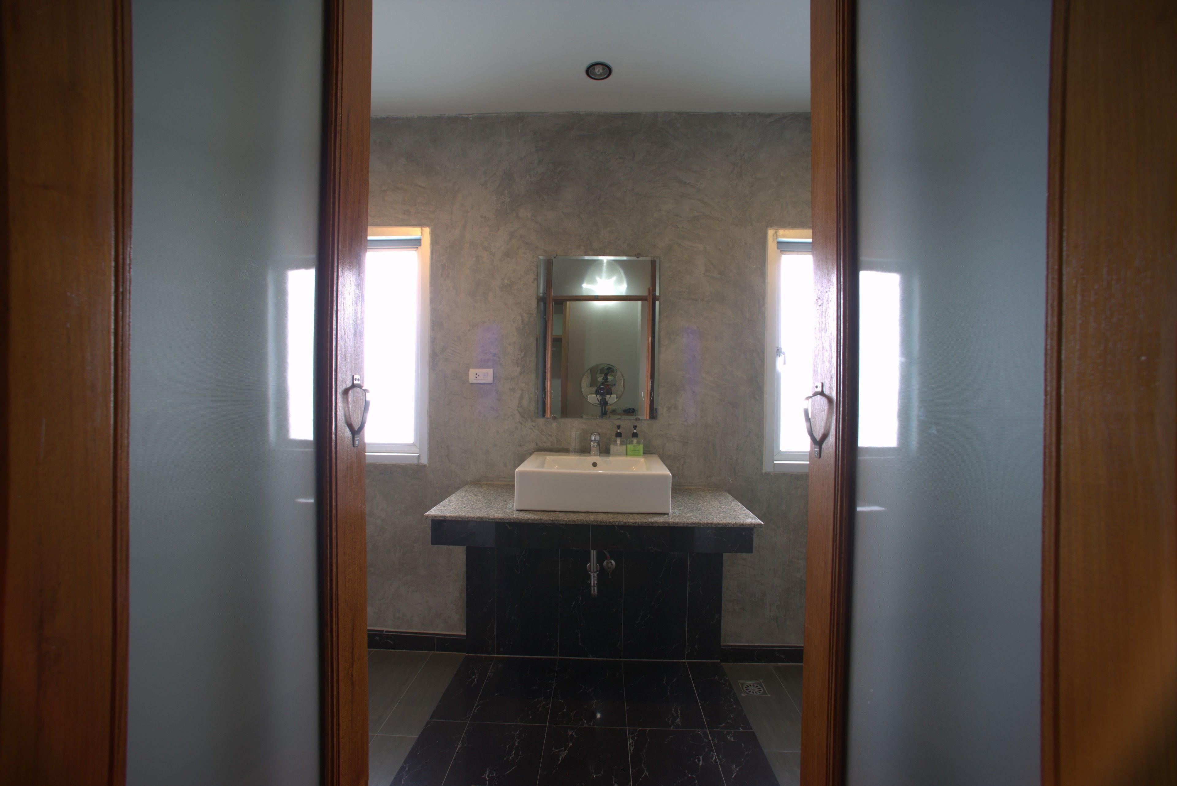 Un baño con un espejo grande, un lavamanos y dos ventanas.