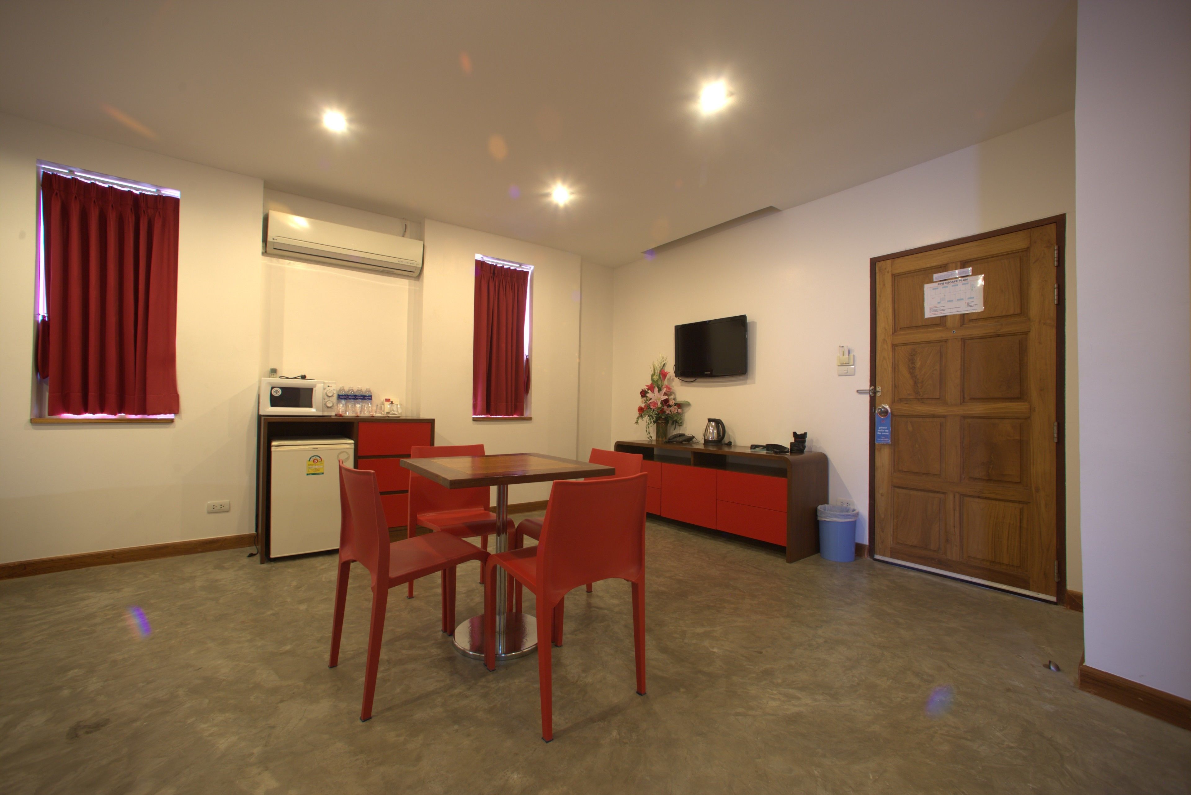 Une chambre d’hôtel compacte dotée d’une kitchenette, d’un coin repas avec des chaises rouges, d’un téléviseur fixé au mur et d’une porte en bois avec une pancarte.