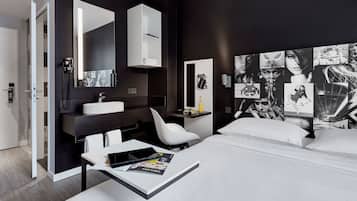 A modern hotel room with a large bed, a bedside table with a magazine, a white chair, a vanity with a mirror and sink, and a wall with black and white photographs.