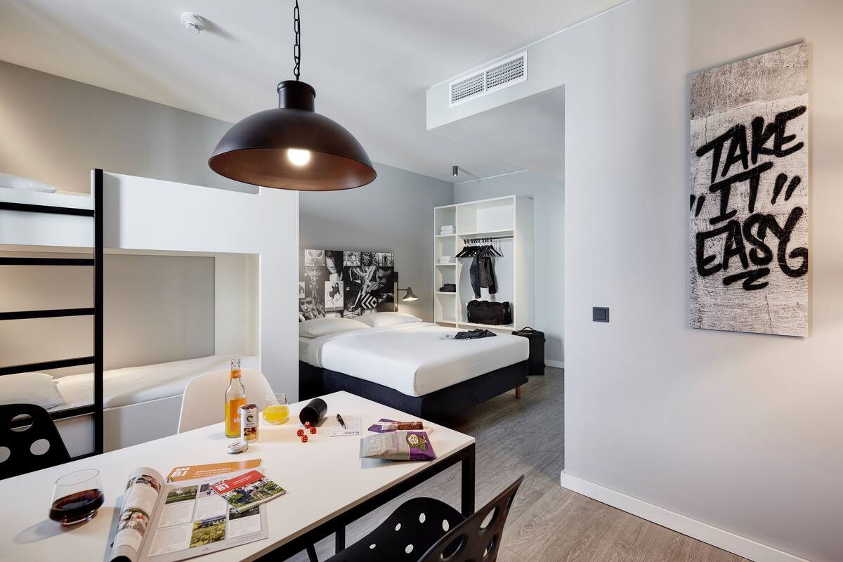 Una habitació d'hotel moderna amb un llit, un escriptori amb una revista, una ampolla i un plat de fruita.