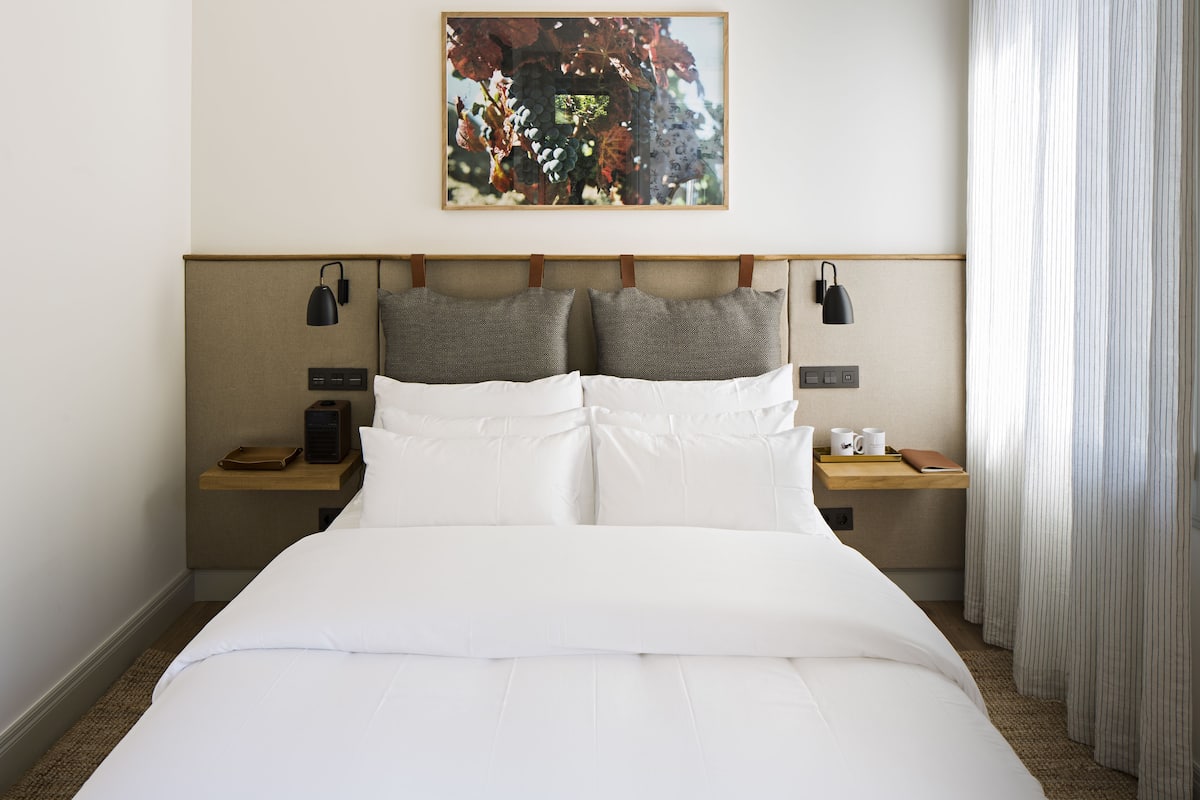 A neatly made bed with white linens, two bedside tables with lamps, a framed picture above the headboard, and a window with curtains.