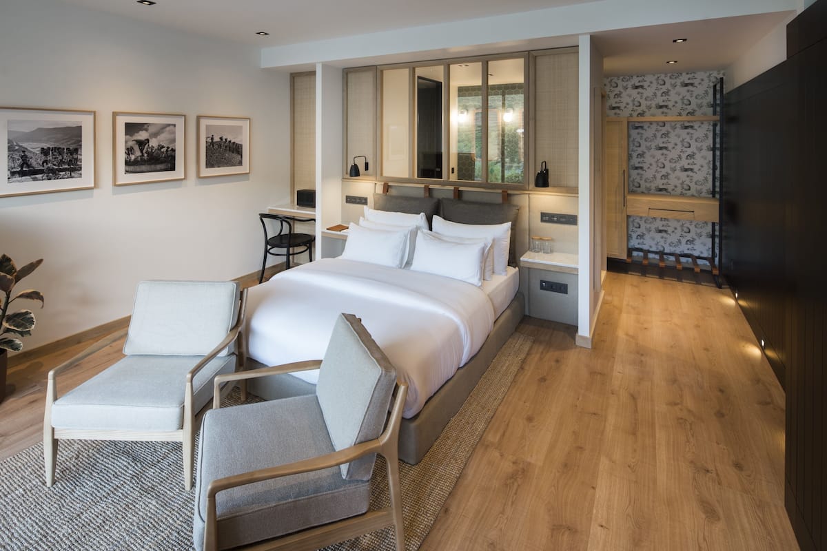 A modern hotel room with a large bed, two bedside tables, a desk, and a chair. There are framed pictures on the wall and a large mirror above the bed.