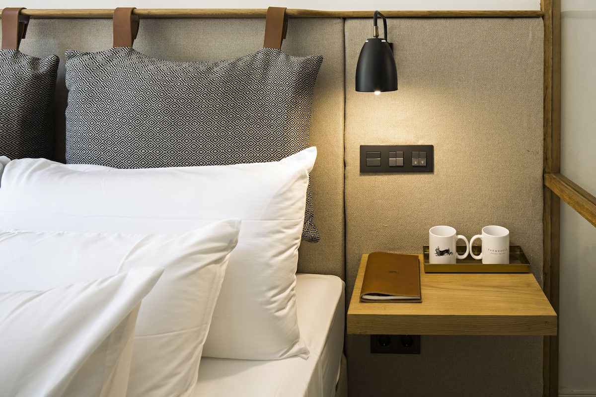 A neatly made bed with white linens and patterned pillows, a bedside table with a lamp, two mugs, and a notebook.