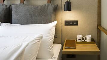 Una cama bien hecha con sábanas blancas y almohadas estampadas, una mesita de noche con lámpara, dos tazas y un cuaderno.