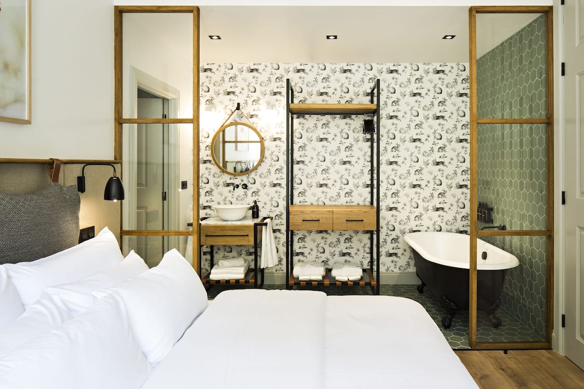 A bedroom with a large bed, a wooden bedside table, a round mirror, a built-in sink, and a freestanding bathtub.