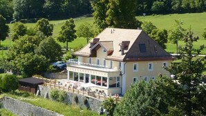 Une grande maison à deux étages dotée d’un balcon, entourée de verdure et d’un mur de pierre.