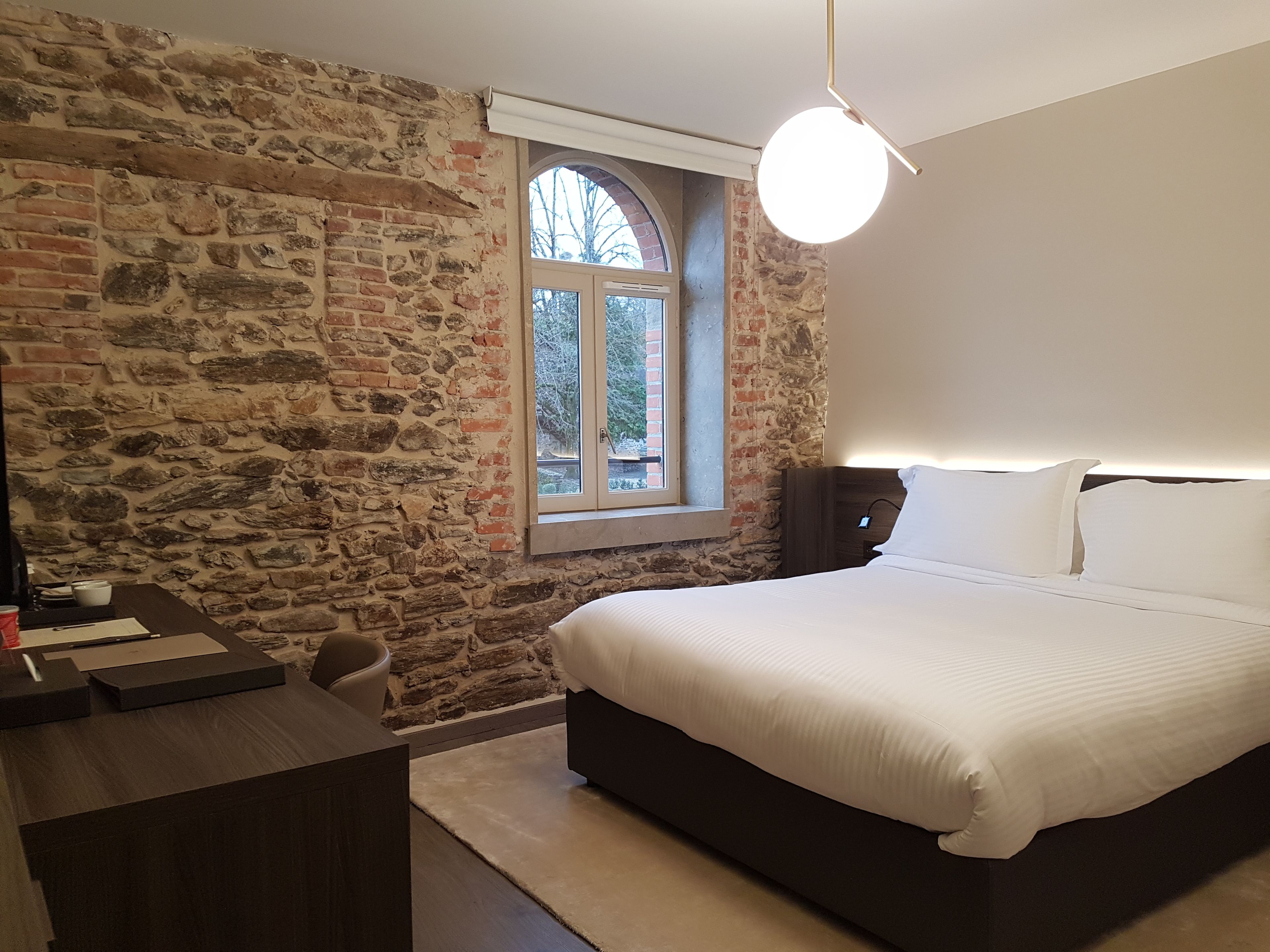 Une chambre avec un mur en pierre, un grand lit, un bureau et une fenêtre donnant sur les arbres.